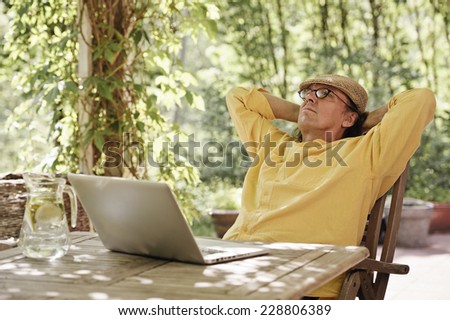 Senior man sits outdoors at a wooden table under an arbor and works on a laptop computer. It\'s summer and there\'s a background of green trees and bushes
