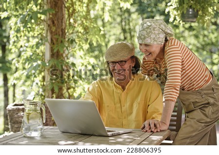 Senior couple outdoors with a laptop, They\'re looking at the computer. Possibly having a wireless video call with grandchildren. Dappled sunlight from the overhead arbor filters through.