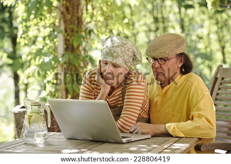 Senior couple outdoors with a laptop, They\'re looking at the computer. There\'s a sunny background of trees and bushes. Dappled sunlight from the arbor overhead filters through