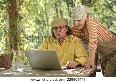 Senior couple outdoors with a laptop, They\'re looking at the computer. Possibly having a wireless video call with grandchildren. Dappled sunlight from the overhead arbor filters through.