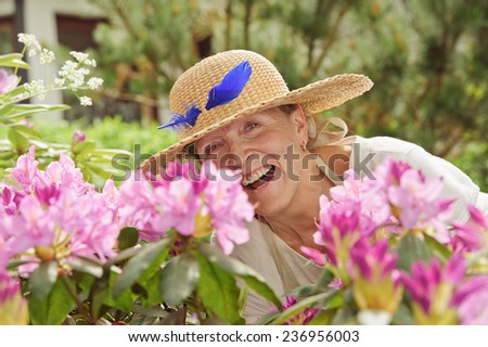 Senior woman laughing behind rhododendron flowers. She\'s wearing a straw hat with a blue feather in it.