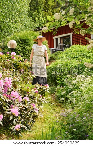Senior woman standing in her garden, She is surrounded by bushes. There\'s a red house partly seen in the background. She\'s wearing an apron