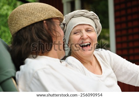 Senior man and woman sitting outdoors. The woman laughs at something the man has said. Window and red wall in the background