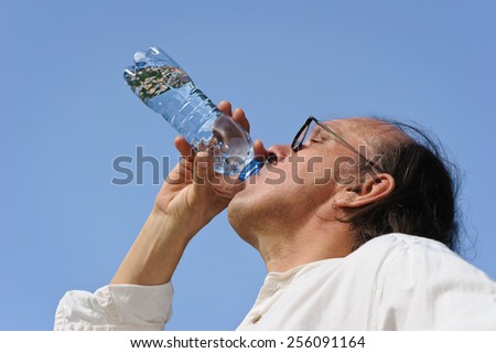 Thirsty senior man drinks water from a bottle outdoors. There is a lake and forest in the background. Low camera angle.