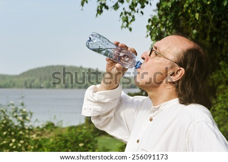 Thirsty senior man drinks water from a bottle outdoors. There is a lake and forest in the background. Digital filter has been used