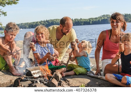 Family with four children grilling sausages over a camp fire beside a lake. The sausages are stuck onto wooden steaks gathered from the forest.
