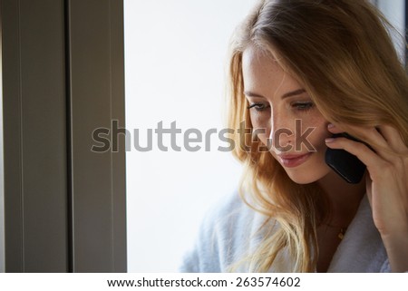 Young businesswoman talking on mobile phone while standing by window in office. Beautiful young female model in bright office.