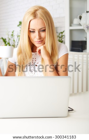 Cute woman having a cup of tea while using her laptop in her kitchen