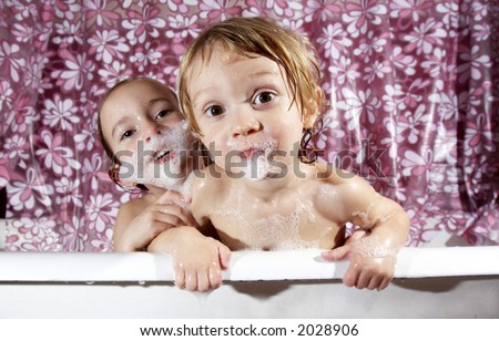 two small young kids play in a bathtub, soapy bubbles on their faces, the little one holding onto the edge of the bath tub with water dripping. A colorful shower curtain with flowers is the backdrop.