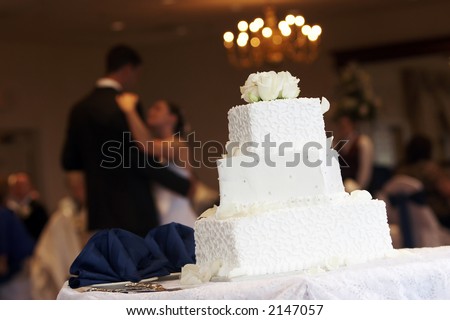 a white wedding cake with neat little swirl details and silver candy buttons, bride and groom are dancing in the background