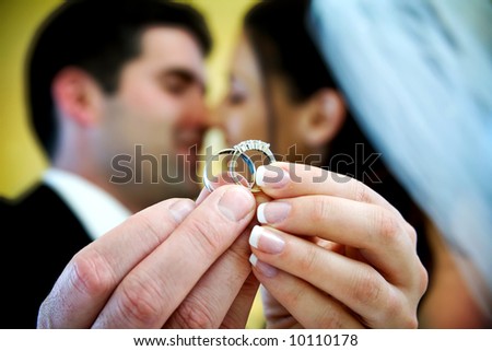 The bride and groom are holding up the wedding rings and kissing in the background. NOTE: this photo has a very shallow depth of field
