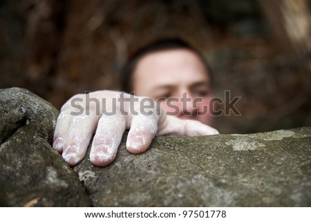 A climber reaches the top of a boulder. His visible hand is covered in white chalk, and his face is just peeking over the edge.
