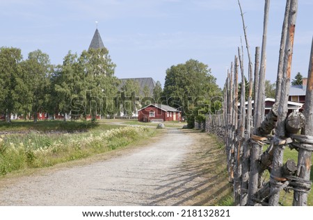 A traditionally wooden fence this side a village which forms the boundary line. Buildings and church in the background. A gravel road leads to the village.