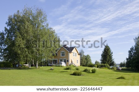 Yellow wooden house in the Nordic countryside. Garden, park this side, trees ans sunshine. Blue sky.