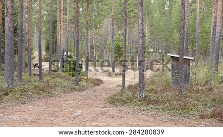 Campsite in a forest with table and outhouse. High pines, bushy and a black car. Path into the area.