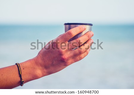 Woman\'s hand holding a thermos cup by the sea