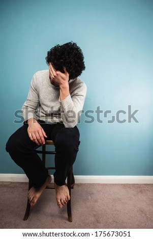 A sad young man is sitting on a high chair by a blue wall