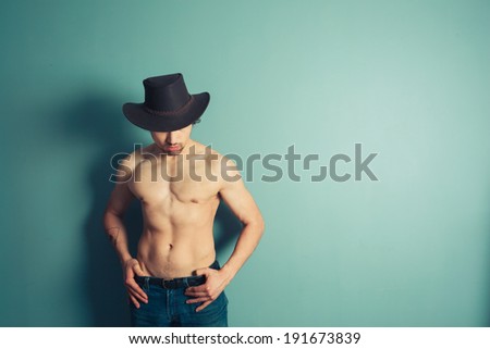 A sexy shirtless cowboy is posing by a blue wall
