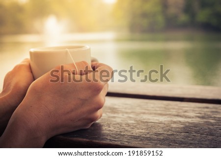Closeup on a woman\'s hands holding a cup of tea by a lake in the afternoon