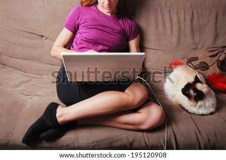 A young woman is sitting on a sofa and using a laptop computer with a cat sitting next to her