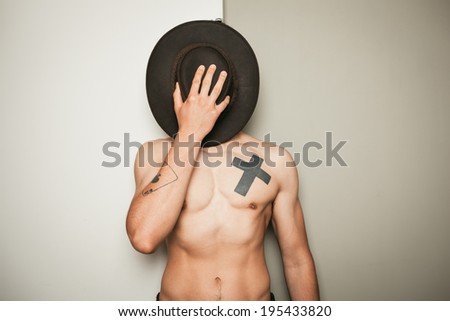 A young shirtless man is hiding his face behind a cowboy hat