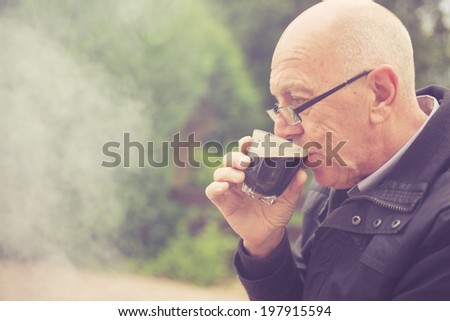 An old man is drinking in a garden with smoke from a barbecue in the background