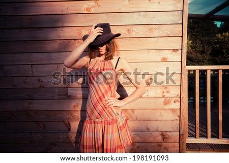 A young woman wearing a cowboy hat is standing outside a wooden cabin