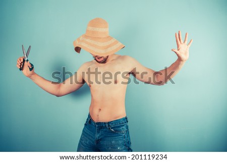 A silly young man wearing a beach hat is playing with scissors