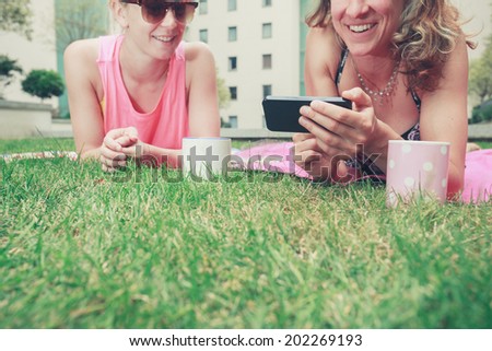 Two young women are laughing and smiling as they look at a phone outside