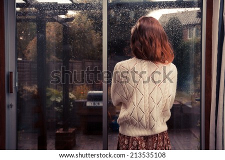 A young woman is standing by the french doors in her house and is looking out at the rain