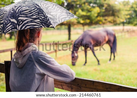 A young woman with an umbrella is standing by a fence and is looking at a horse
