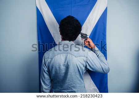 A man is standing in front of a Scottish flag and is pointing a gun to his head to commit suicide