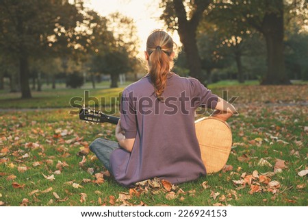 Rear view of young woman sitting on the grass and playing guitar in the park at sunset