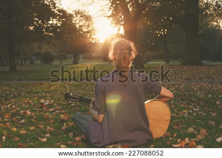 Rear view of young woman sitting on the grass and playing guitar in the park at sunset