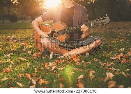 A young woman is sitting on the grass in the park and is playing guitar at sunset