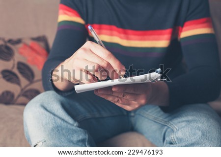 A young man is sitting on a sofa and is writing on the back of an envelope