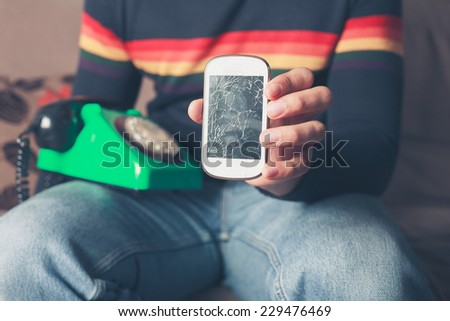A young man is sitting on a sofa with a broken smart phone and an old rotary phone