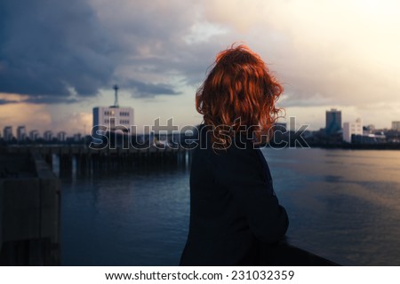 A young woman is admiring the sunset over a river in the city on a cold autumn day