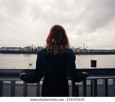 A young woman is standing on a wooden terrace by the river with a glass of beer