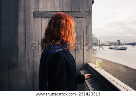 A young woman is standing on a wooden terrace by the river with a glass of beer