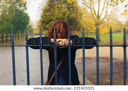 A sad woman is resting her head on a gate in the park in autumn