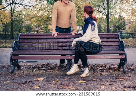 A young woman is sitting on a park bench and is talking to a man who is standing behind her