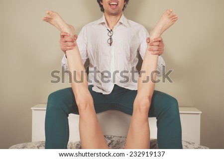 A happy young man is sitting on the headboard of a bed and is spreading a woman\'s legs