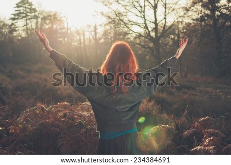 A young woman is raising her arms in joy as she is standing in a forest at sunset