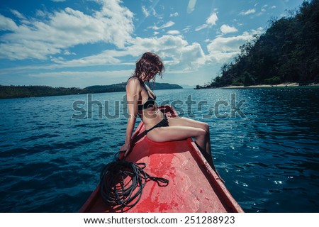 A sexy young woman wearing a bikini is sitting on a small boat near a tropical island