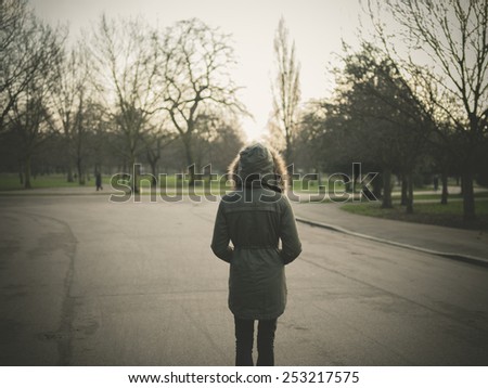 A person wearing a warm coat with a big hood is standing in a park at sunset