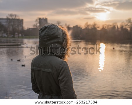 A person wearing a thick warm coat is standing by a pond in a  park and is watching the sunset in winter