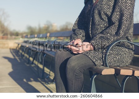 A young woman is sitting on a bench by a water feature in a park and is using her phone