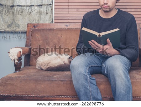 A young man is sitting on a sofa with a cat and is reading a big book
