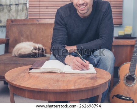 A young man is sitting with his cat and guitar at home on a sofa and is writing songs in a notebook